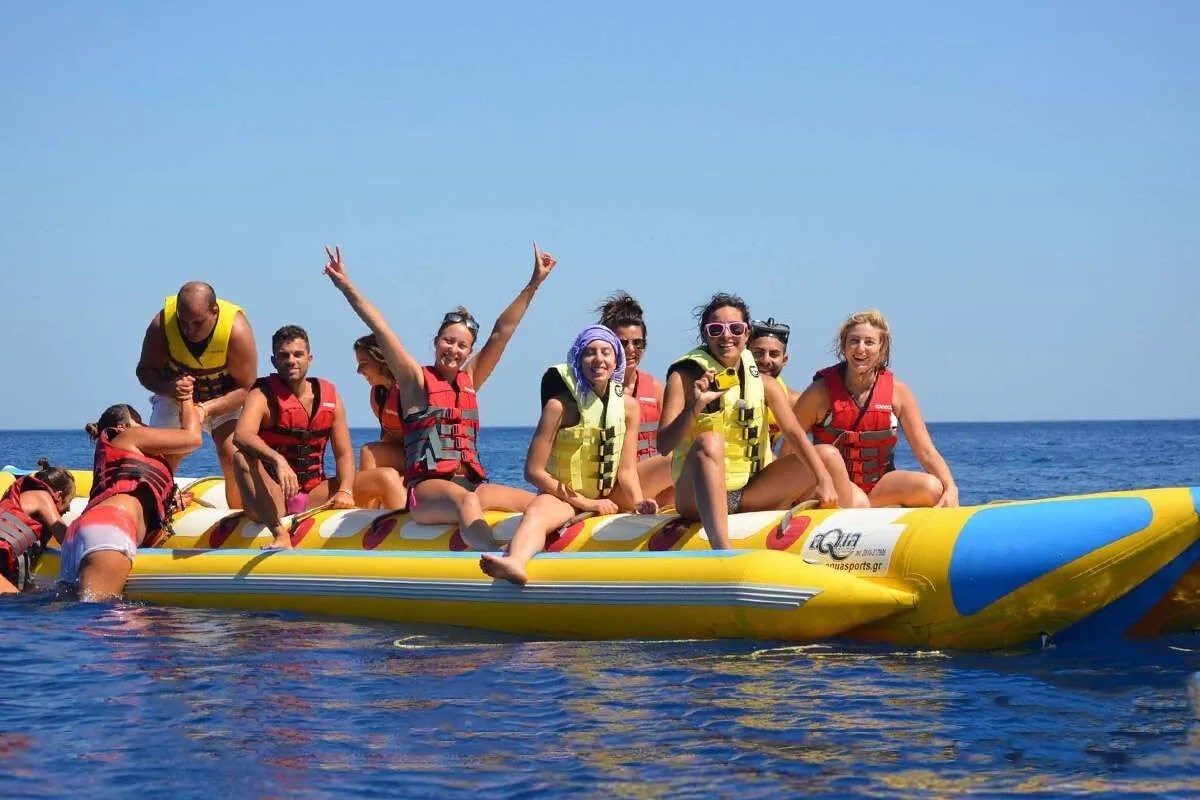 Group of people on an inflatable banana boat, wearing life vests, smiling and waving on a sunny day.