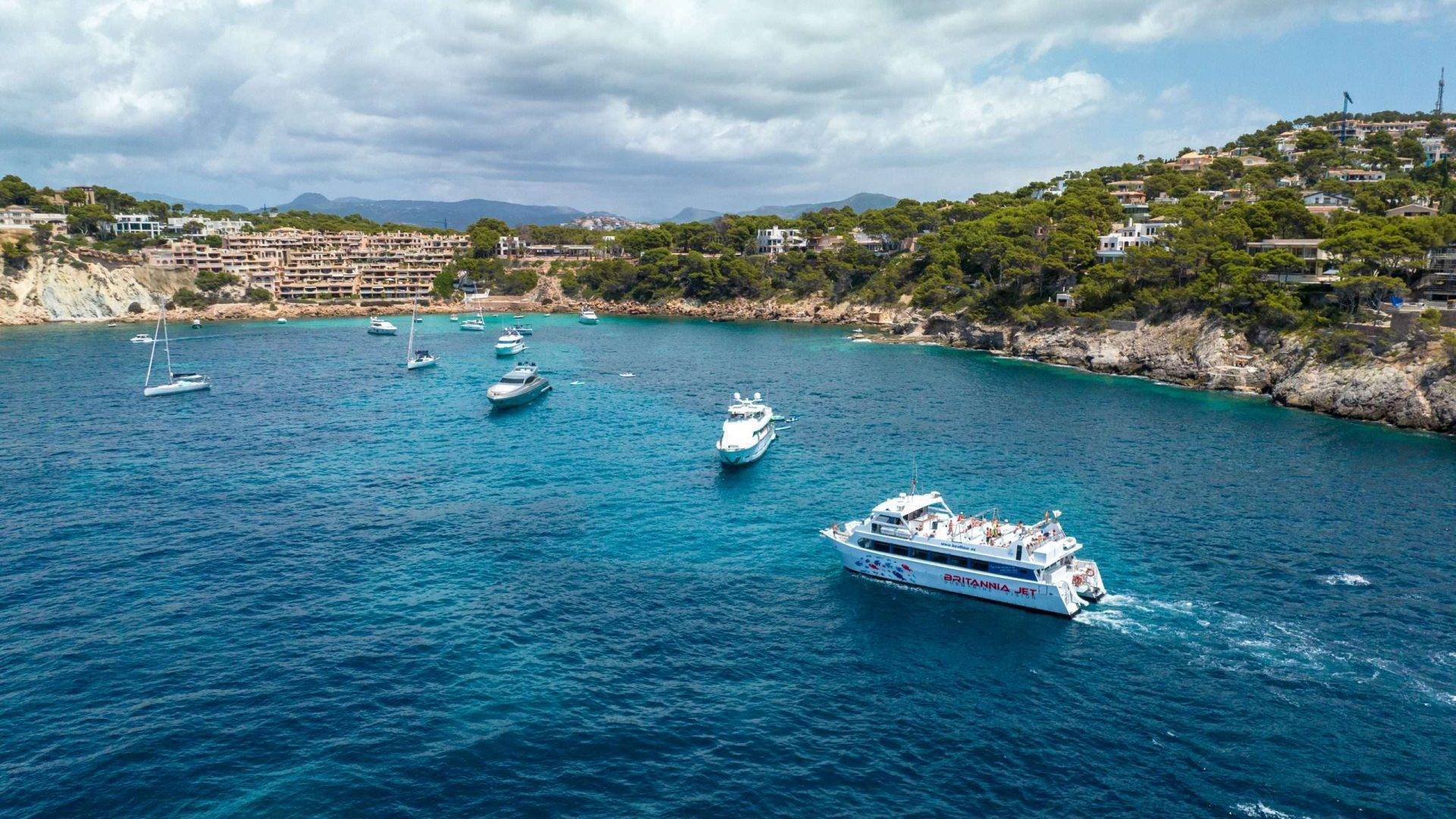 Boats in a scenic bay with clear blue water, rocky cliffs, and buildings on the coastline.