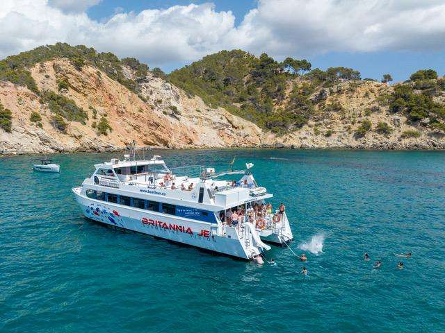 Large boat near a rocky coastline with swimmers nearby on a sunny day.