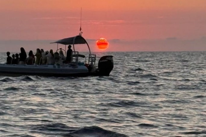 Boat with people on ocean at sunset, sun near horizon.