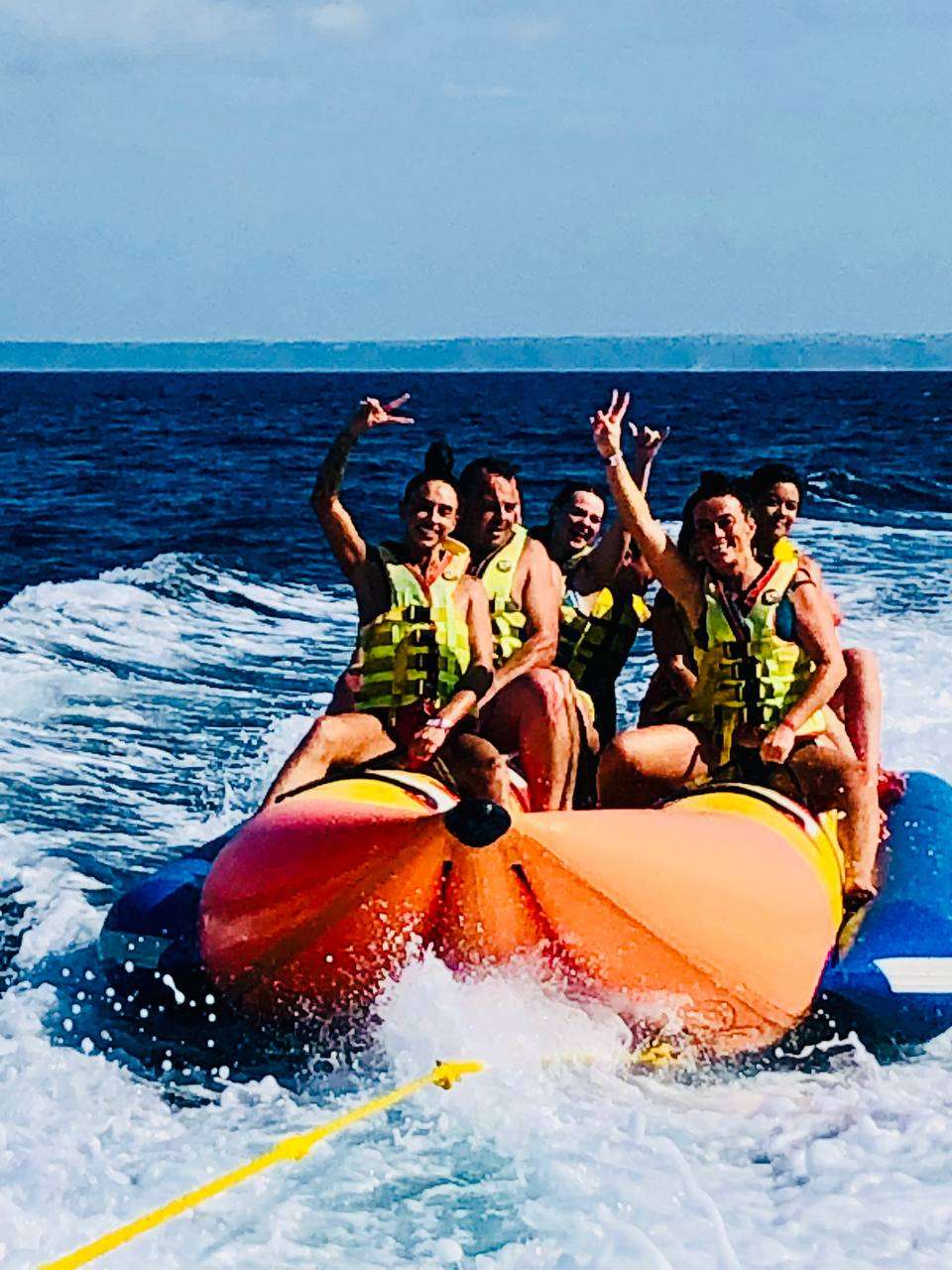 Group of people on an inflatable banana boat in the ocean, wearing life jackets and making hand gestures.