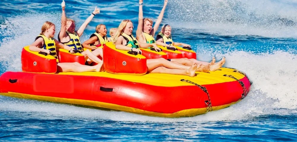 Group of people on a colorful inflatable raft in the ocean, laughing and raising arms.