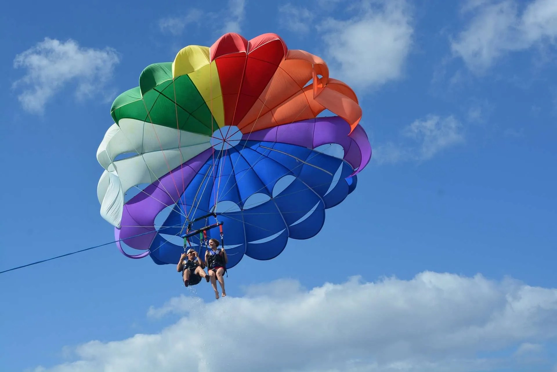 Two people parasailing with a rainbow-colored parachute against a blue sky.
