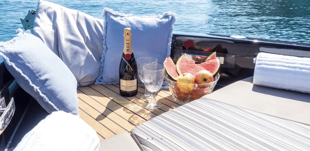 Luxury boat deck with cushions, champagne, glass, and fruit bowl overlooking water.