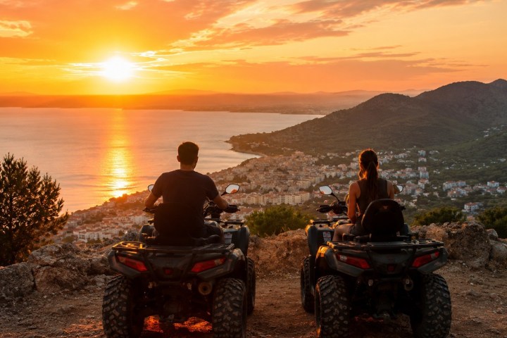 Two people on ATVs watching a coastal sunset from a hilltop.
